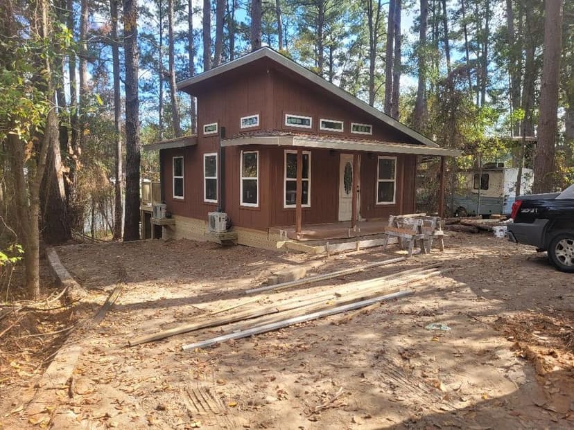 Two-story wooden cabin surrounded by tall trees, with a porch and construction materials outside.